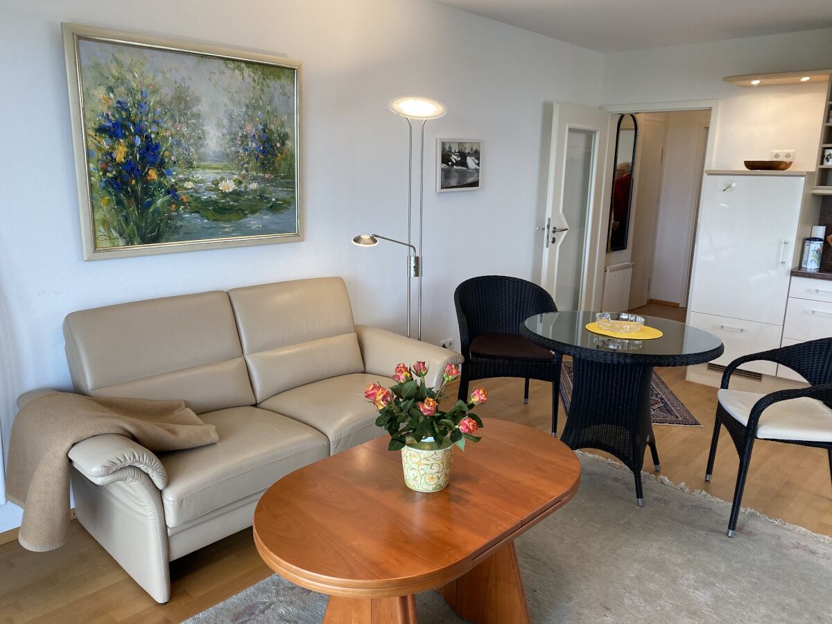 Ett vardagsrum med en beige soffa, ett träbord med en vas med blommor, ett matbord med stolar och en tavla på väggen.