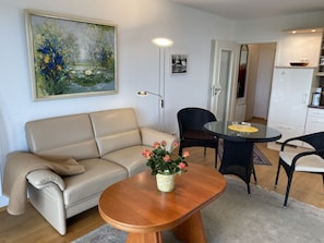 A living room with a beige sofa, a wooden coffee table with a vase of flowers, a dining table with chairs, and a painting on the wall.
