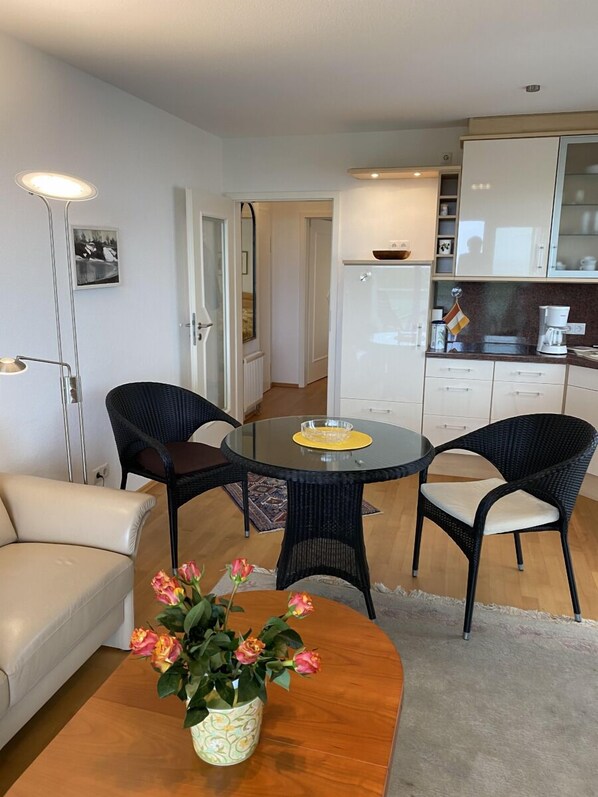 A compact living space with a round glass table, two black wicker chairs, a beige sofa, and a small wooden coffee table with a vase of flowers. A kitchen with white cabinets and a refrigerator is visible in the background.
