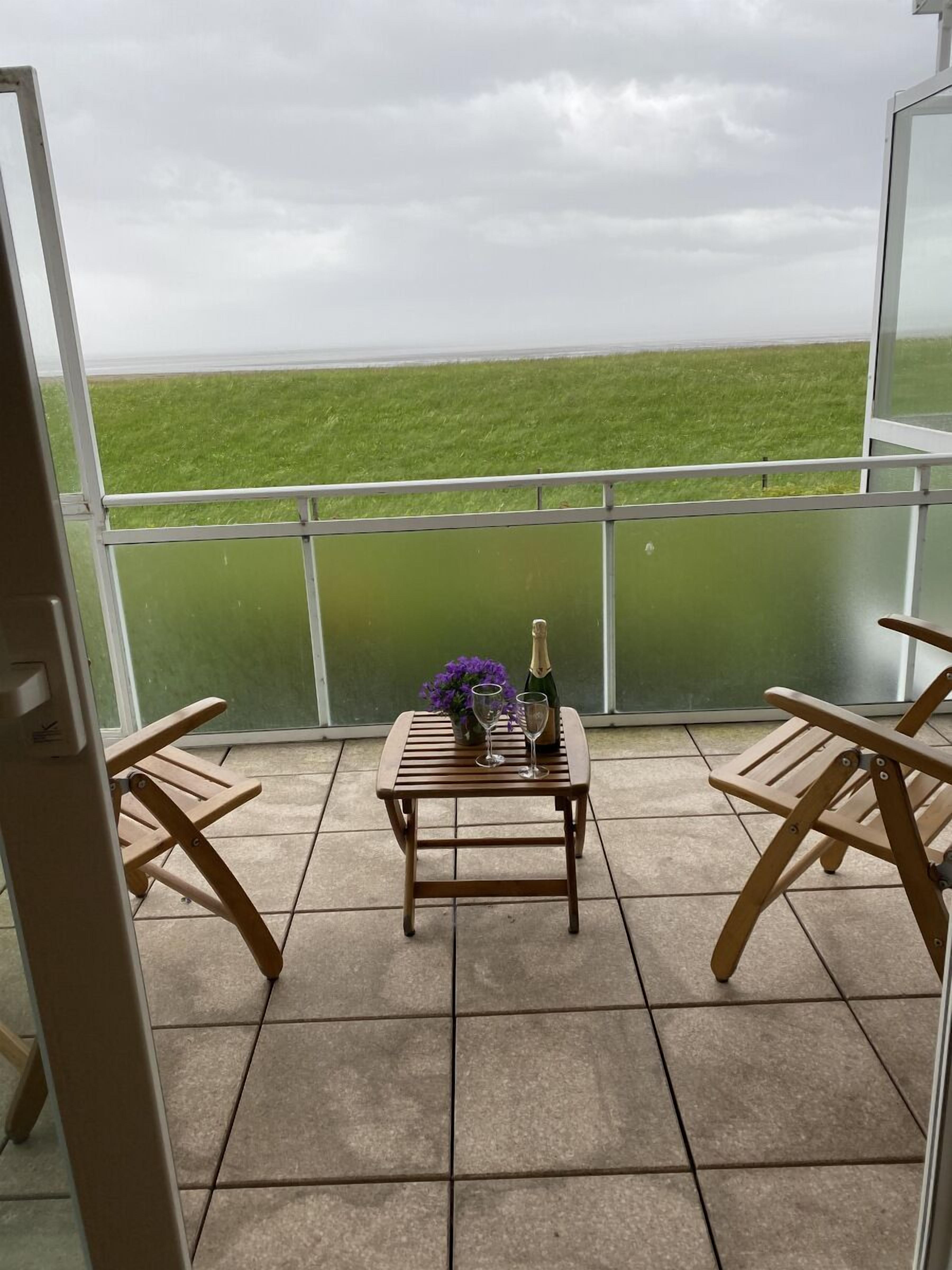 Varanda com vista para um campo verde, mesa com garrafa e copos, e duas cadeiras de madeira.