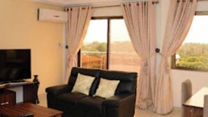 Ein Wohnzimmer mit einem schwarzen Sofa, einem Holztisch, einem Fernseher und Blick ins Freie durch große Fenster.