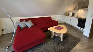 Ein gemütlicher, kleiner Wohnraum mit einem roten Sofa, einem kleinen Holztisch und einer Küchenzeile mit weißen Schränken und einem Herd.