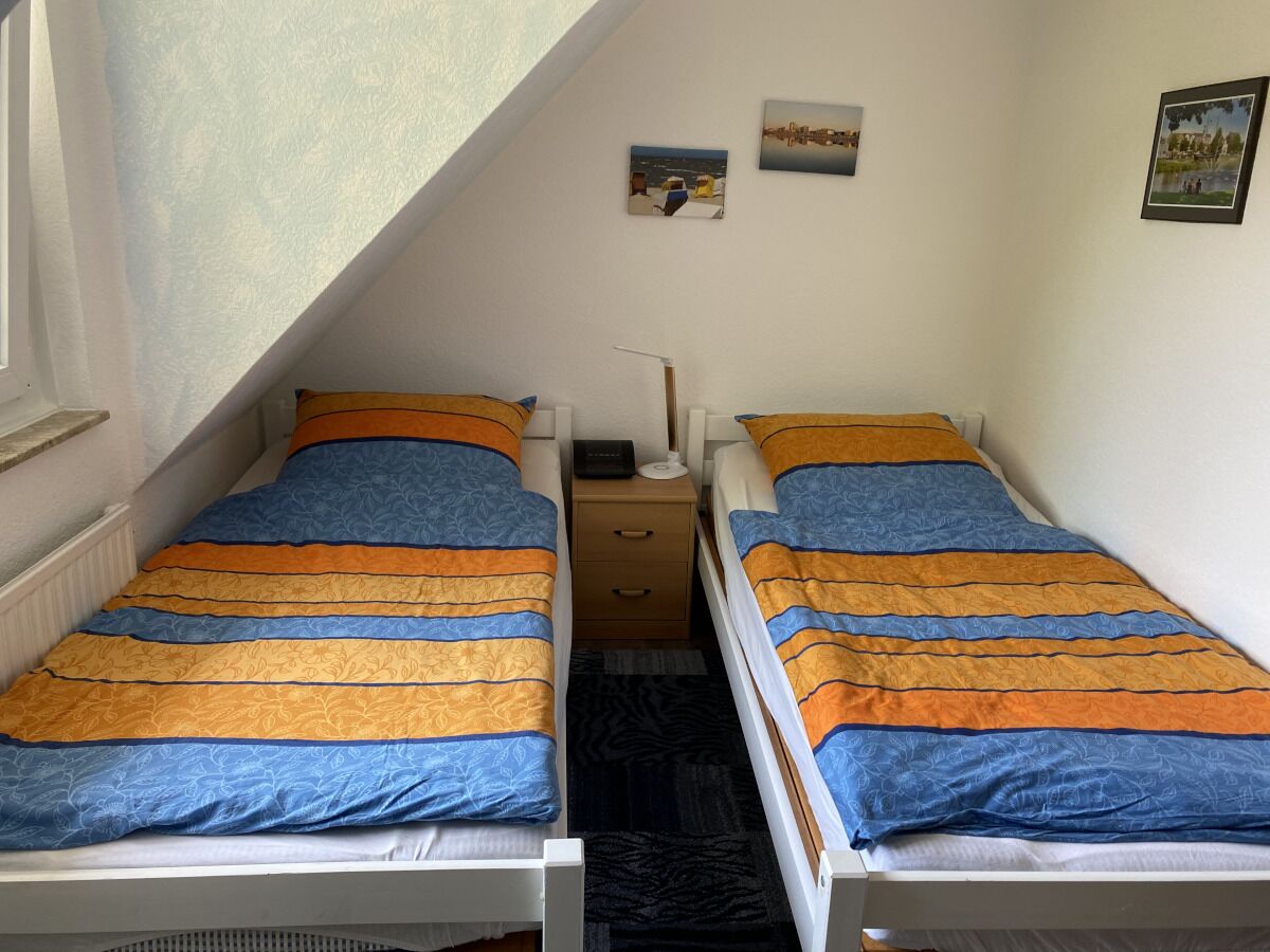 Una habitación tipo dormitorio con dos camas, cada una con una manta a rayas. Hay una mesita de noche pequeña con una lámpara y un cajón. La habitación tiene un techo inclinado y una ventana en el lado izquierdo.