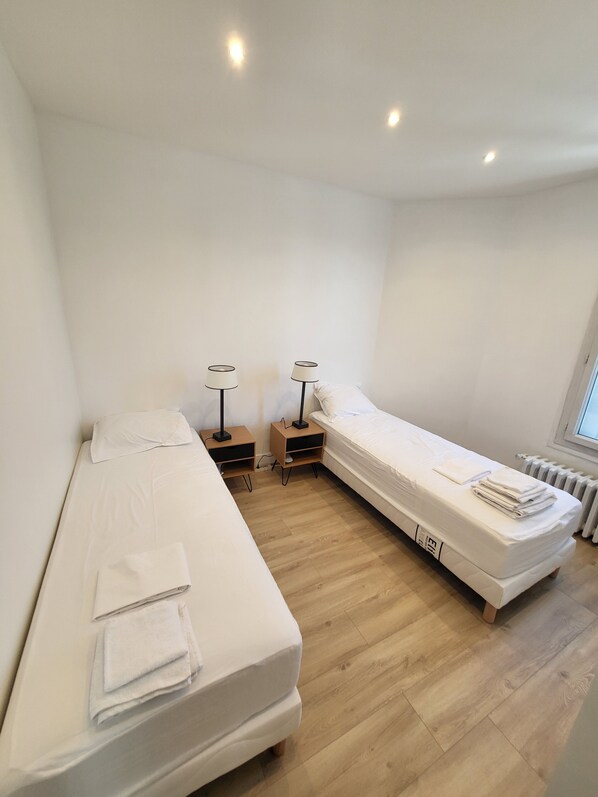 A small, well-lit room with two single beds, a nightstand with a lamp, and a radiator.