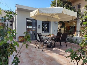 Une terrasse extérieure avec une table et des chaises sous un grand parasol, entourée de plantes en pot et d’un bâtiment doté d’une porte-fenêtre coulissante.