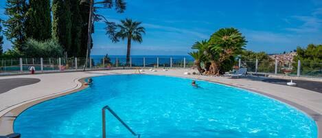 A large, clear blue pool with a curved edge, surrounded by a paved deck and a metal fence. There are palm trees and other greenery around the pool area.