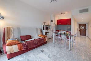 A modern living room with a sofa, a glass dining table, and a kitchen area with red cabinets. - Appartement Menton Garavan Avec Superbe Piscine vue mer Proche Italie & Monaco (Menton)