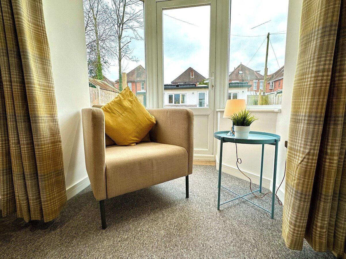 A room with a beige armchair, a small table with a plant, and a view of houses through a window.