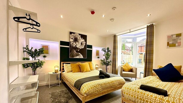 A modern hotel room with a large bed, a smaller bed, a desk, and a chair. There are plants and artwork on the walls. - Massive New House Sleeps 20+ Accepts Groups (Bournemouth)