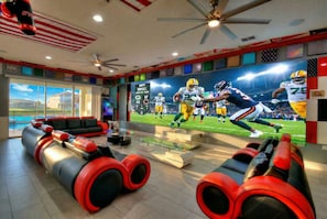 A room with a large TV screen displaying a football game, a ceiling fan, and a seating area with red and black couches. - 2 Side-by-side homes w Bowling, 1GB wifi, videowall TVs, ensuites, sleep 80 (Kissimmee)