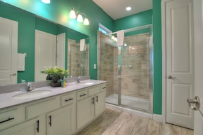 A modern bathroom with a large mirror, two sinks, a shower with glass doors, and a potted plant on the vanity.