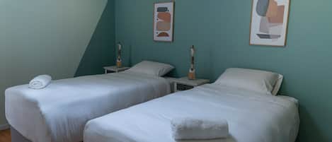 A room with two single beds, each with a white bedspread and pillows, and a small bedside table with a lamp.