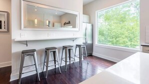 A modern kitchen with a large mirror, three bar stools, and a view of greenery through a large window. - The Taylor (Nashville)