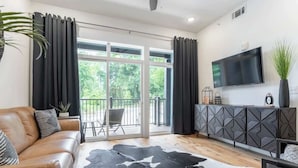 A modern living room with a leather sofa, a black and white cowhide rug, a flat-screen TV mounted on the wall, and a balcony with a view of greenery.
