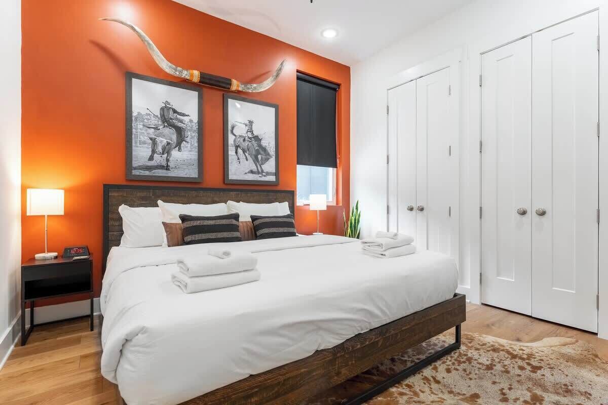 A modern bedroom with a large bed, a wooden headboard, two framed pictures on the wall, a nightstand with a lamp, and a large window with a black blind.