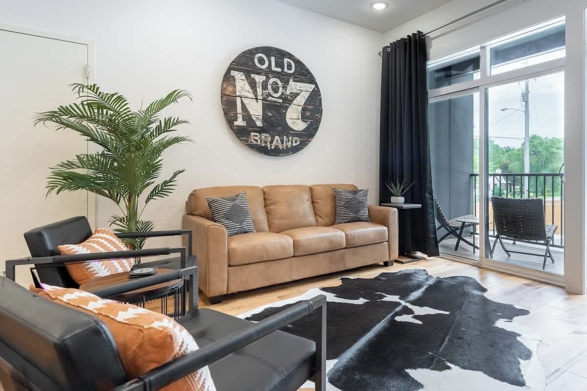 A modern living room with a leather sofa, a black armchair, and a black and white cowhide rug. There is a large window with a view of greenery outside.