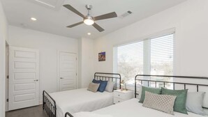 Quarto com duas camas, um ventilador de teto, uma janela com persianas e uma mesa de cabeceira com abajur.