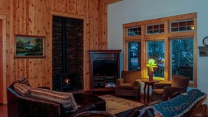 A cozy living room with wooden walls, a fireplace, a television, and a view of the outside through large windows.