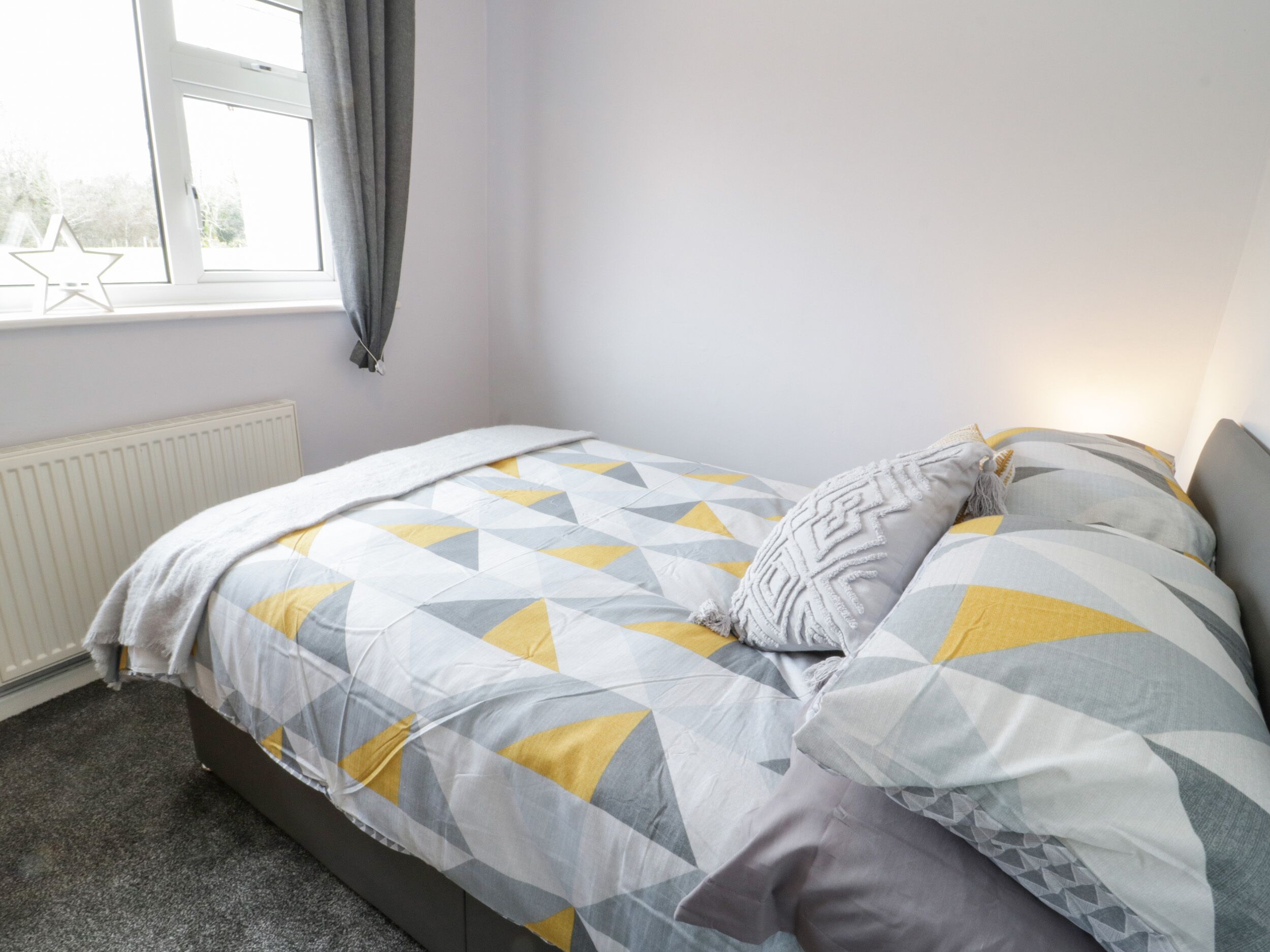 Un dormitorio con cama, ventana con cortinas, radiador y piso cubierto de alfombra.