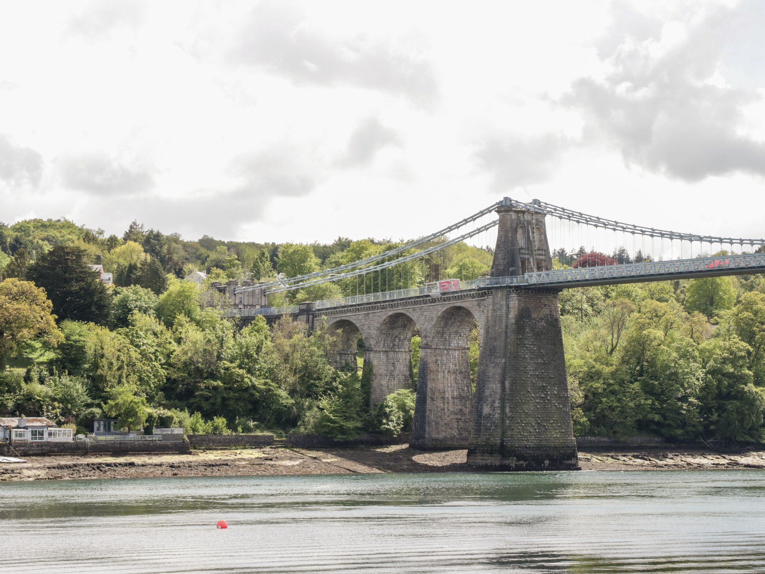 Un pont en pierre doté d’une section suspendue qui enjambe une rivière, entouré d’arbres et avec une colline en arrière-plan.