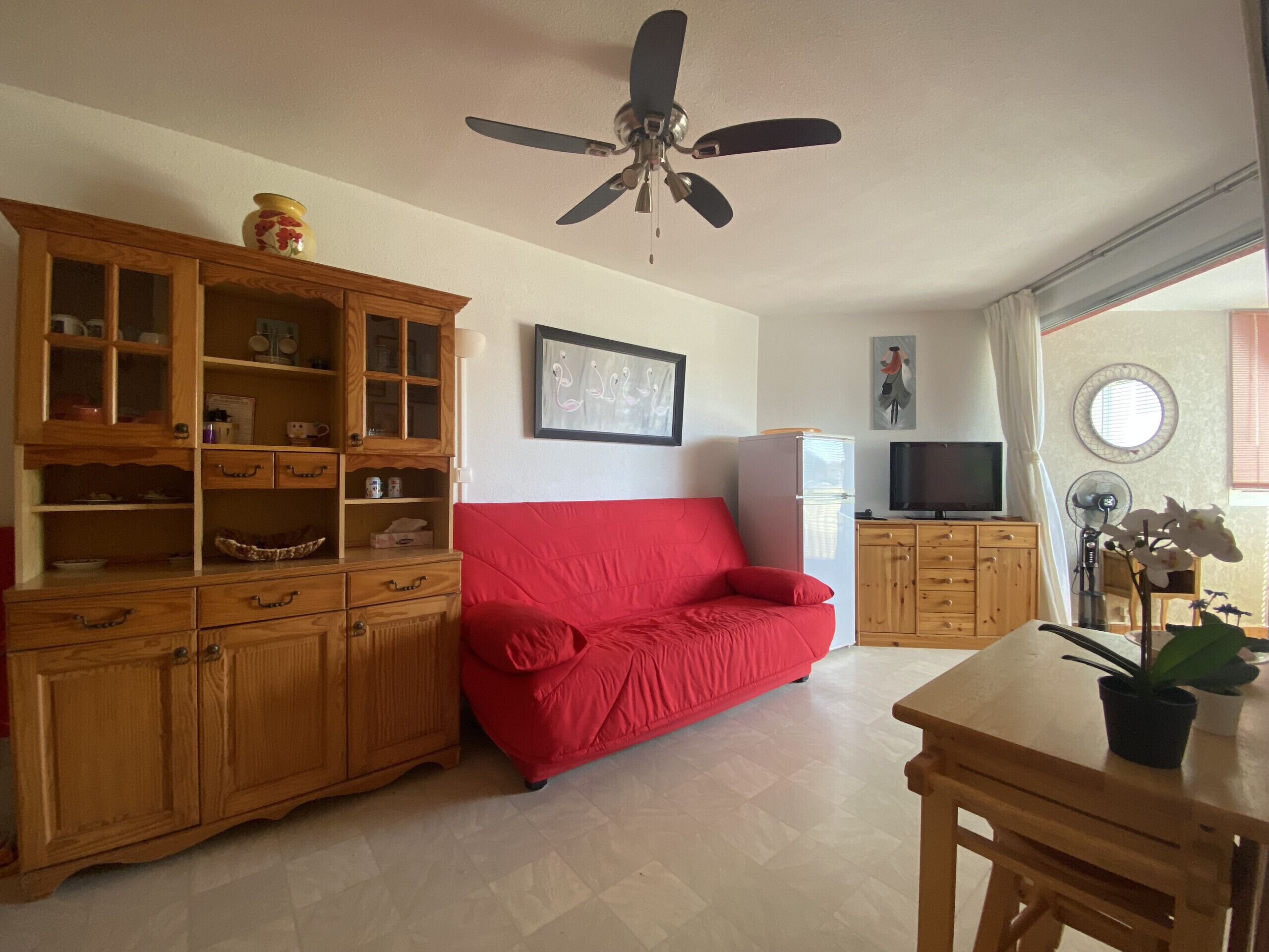 A living room with a red sofa, wooden cabinets, a ceiling fan, a television, and a potted plant.