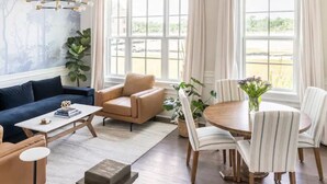 Un salon comprenant un canapé, des fauteuils, une table basse, une table à manger avec des chaises, et de grandes fenêtres avec des rideaux.