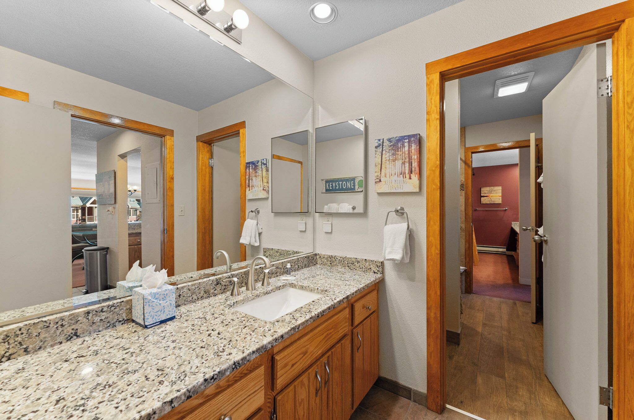 A hotel bathroom with a granite countertop, two sinks, wooden cabinets, a large mirror, and a door leading to another room.