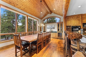 A spacious dining area with a long wooden table and chairs, large windows offering a view of the forest, and a kitchen with wooden cabinets and modern appliances.