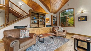 A spacious living room with a wooden staircase, leather sofas, a coffee table, and a large window offering a view of trees. - 140 Goldenrod Circle by Summit County Mountain Retreats (Keystone)