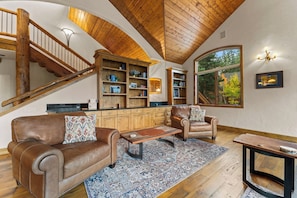 A spacious living room with a wooden staircase, leather sofas, a coffee table, and a large window offering a view of trees.