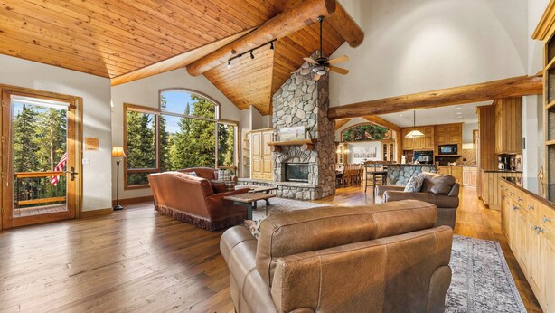 A spacious living area with a stone fireplace, wooden ceiling beams, and a large window overlooking a forest. - 140 Goldenrod Circle by Summit County Mountain Retreats (Keystone)