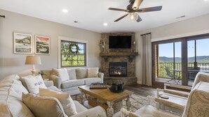 A spacious living room with a stone fireplace, a large TV mounted above it, and a ceiling fan. There are multiple seating areas with sofas and a coffee table.
