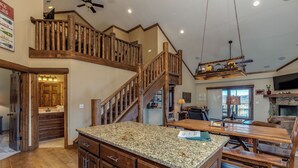 Une cuisine spacieuse dotée de plans de travail en granit, d’armoires en bois et d’un grand îlot central. Un escalier en bois mène à l’étage supérieur, et un lustre est suspendu au plafond.