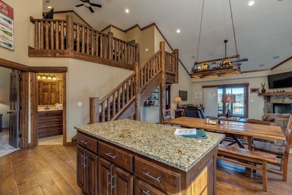 A spacious kitchen with granite countertops, wooden cabinets, and a large island. A wooden staircase leads to an upper level, and a chandelier hangs from the ceiling. - 3BR+Loft Village At Indian Point 1406 near SDC -Free Tickets up to $500 Value (Branson)