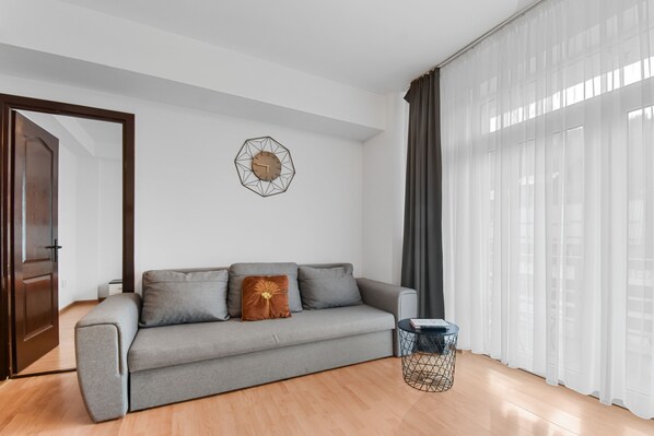 A modern living room with a grey sofa, a geometric wall clock, and a window with sheer curtains. - Pop Apartment (Sinaia)
