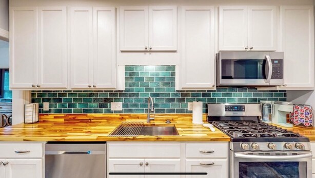 A modern kitchen with white cabinets, a stainless steel stove, a microwave, and a green-tiled backsplash. - Cheerful Four Bedroom home conveniently located (Augusta)