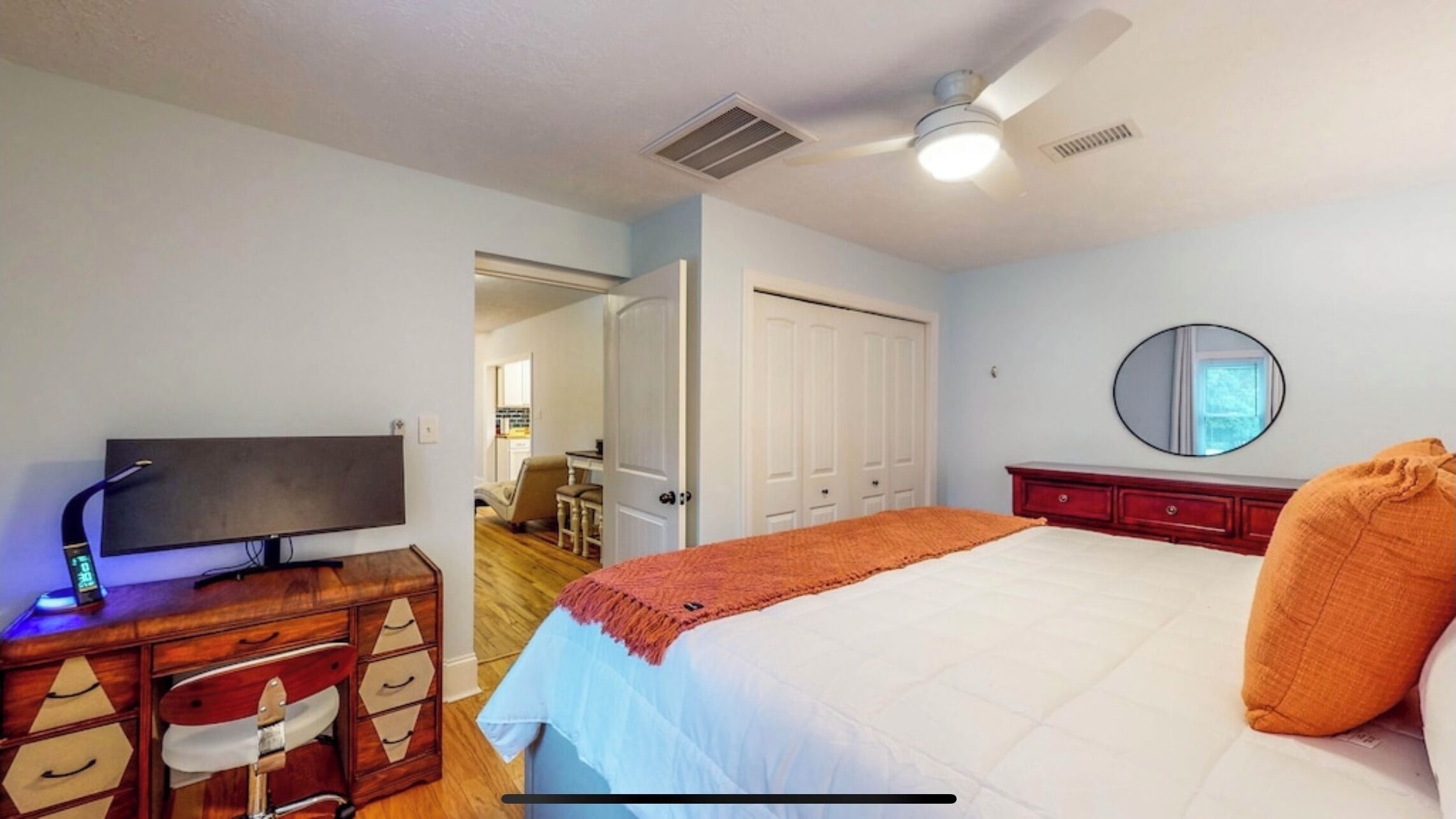 A bedroom with a bed, a television on a wooden dresser, a ceiling fan, and a door leading to another room.