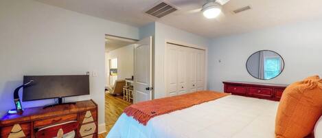 A bedroom with a bed, a television on a wooden dresser, a ceiling fan, and a door leading to another room.