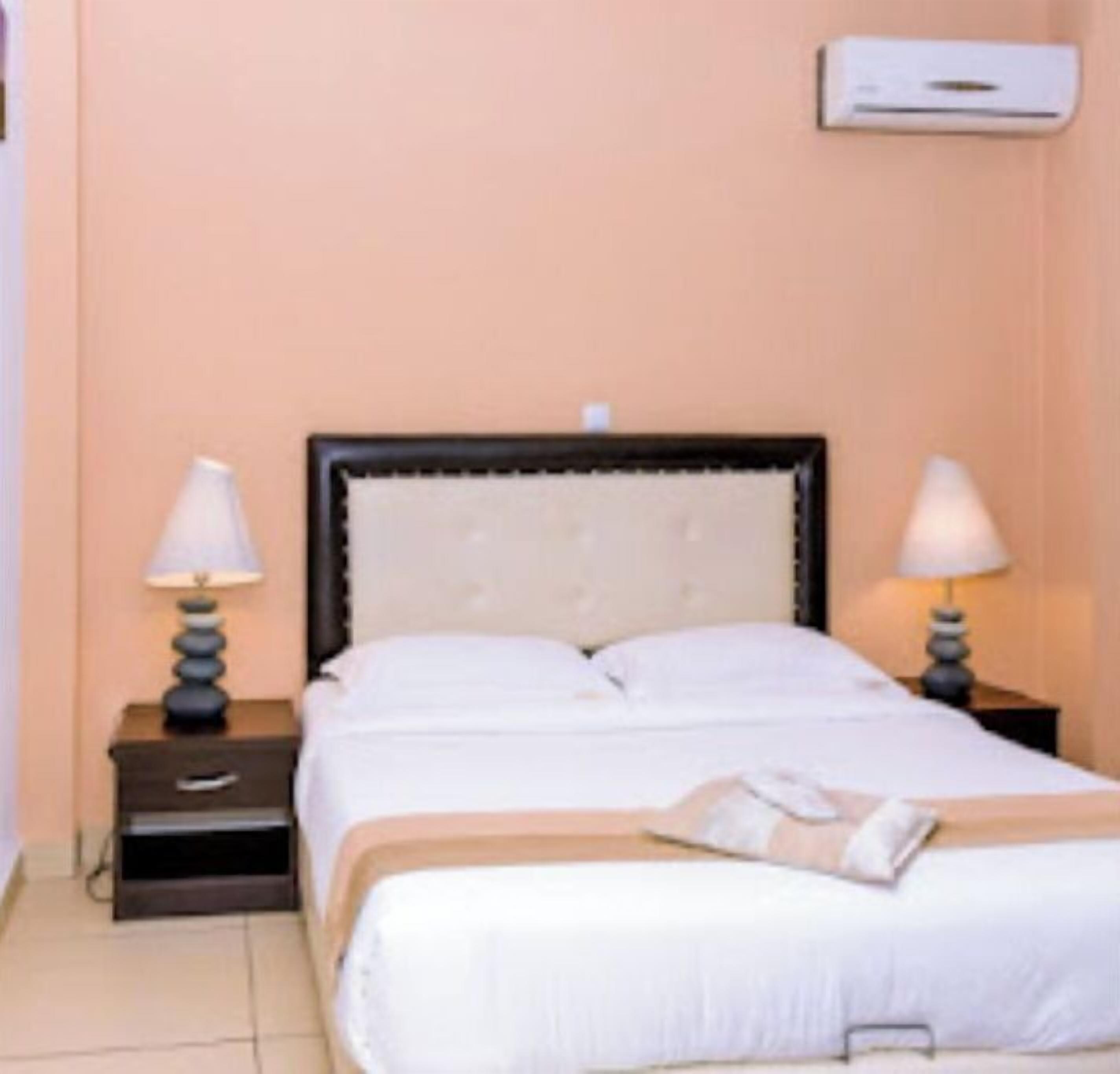 A hotel room with a bed, two bedside tables with lamps, a wall-mounted air conditioner, and a tiled floor.