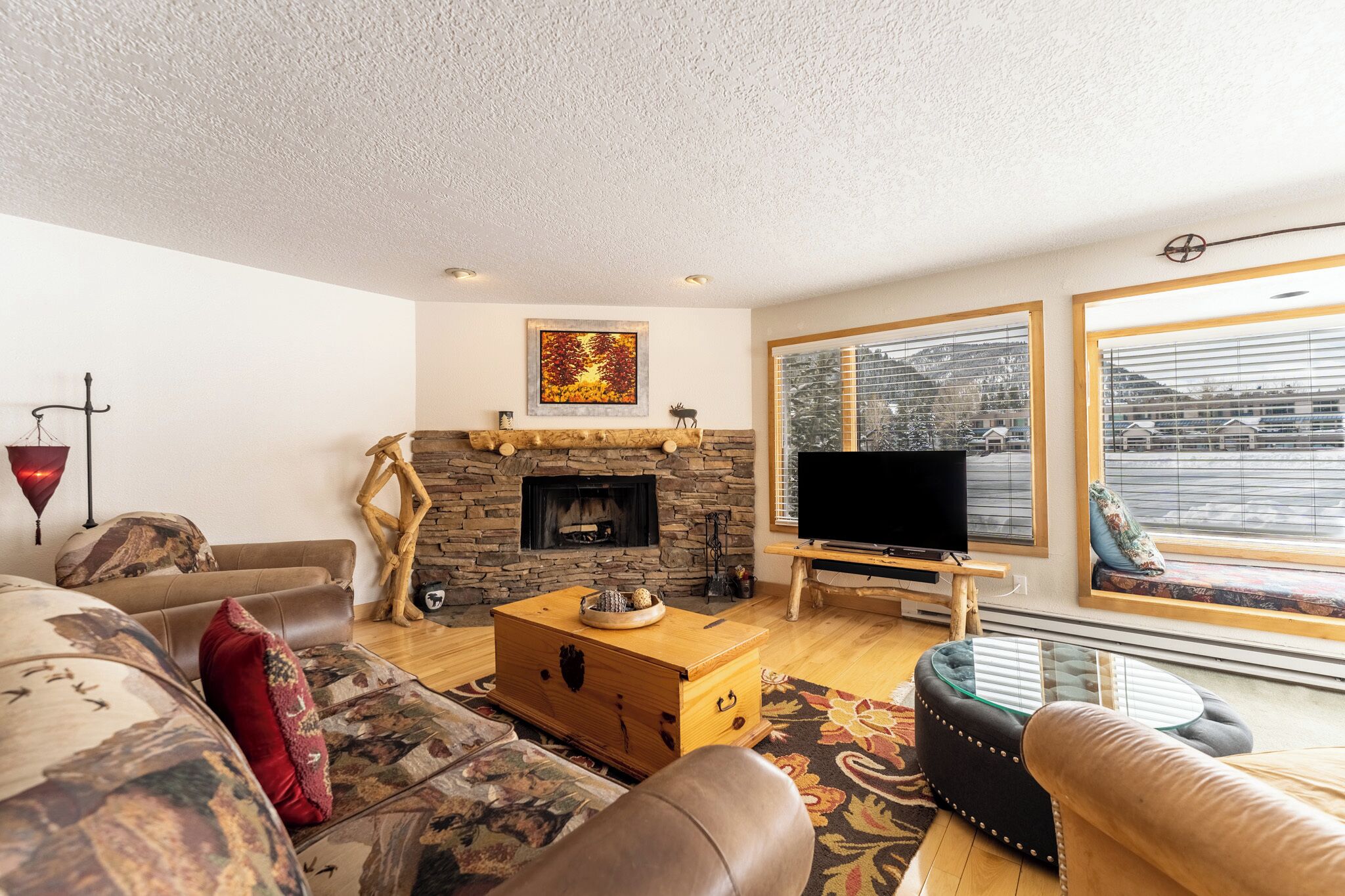 Uma sala de estar com lareira de pedra, televisão e uma janela grande com vista para uma área residencial.
