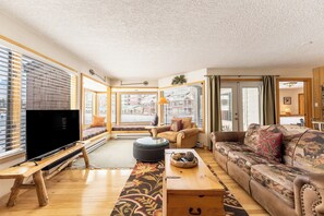 Una sala de estar con un televisor grande, piso de madera, un conjunto de sofá, una mesa de centro y un banco.