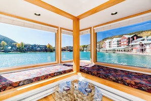 Habitación con amplios ventanales que ofrecen vistas a un lago y edificios. Se observan vigas de madera y una mesa de vidrio con objetos decorativos.