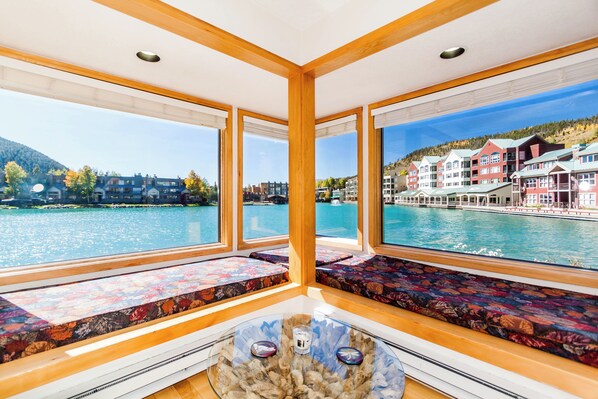 Habitación con amplios ventanales que ofrecen vistas a un lago y edificios. Se observan vigas de madera y una mesa de vidrio con objetos decorativos.