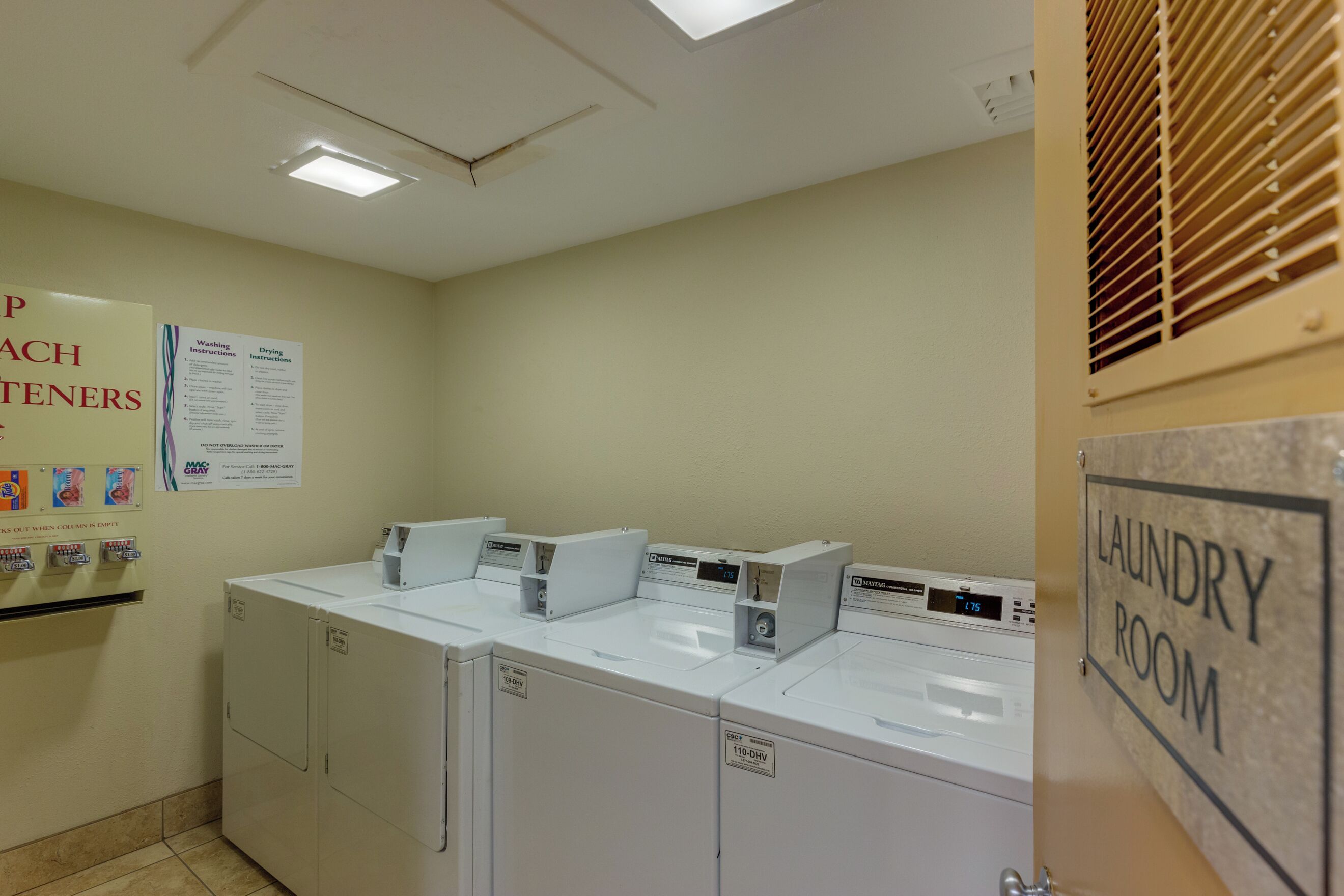 A laundry room with four washing machines, a sign indicating it is a laundry room, and a poster with washing and drying instructions.