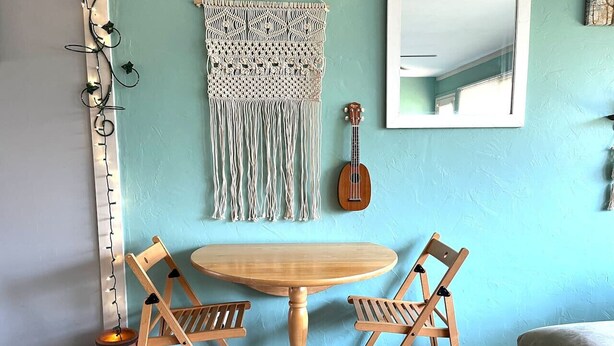 A dining area with a round wooden table and two wooden chairs, a macrame wall hanging, a ukulele, and a large mirror on a light blue wall. - Close to Downtown-Primary on Main-Fast Internet (Greensboro)