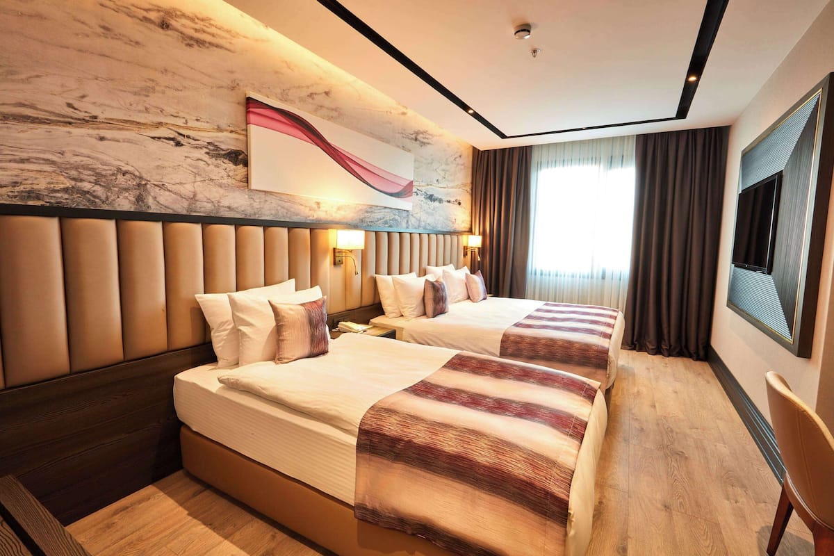 Habitación de hotel con dos camas, pared de mármol, ventana grande con cortinas y televisor de pantalla plana.