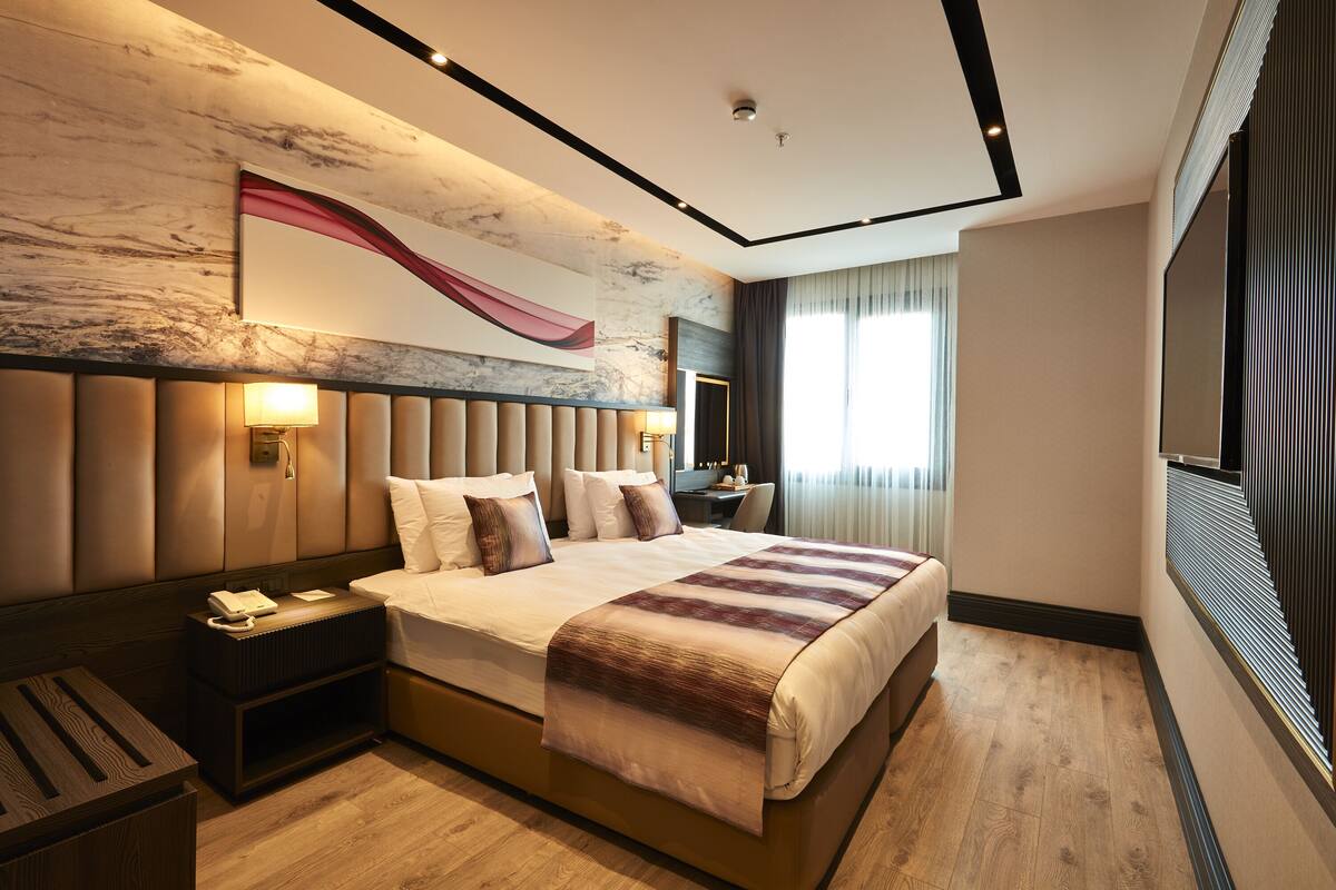 Una habitación de hotel moderna con una cama grande, una mesita de noche con lámpara, un escritorio con silla y un ventanal con cortinas.