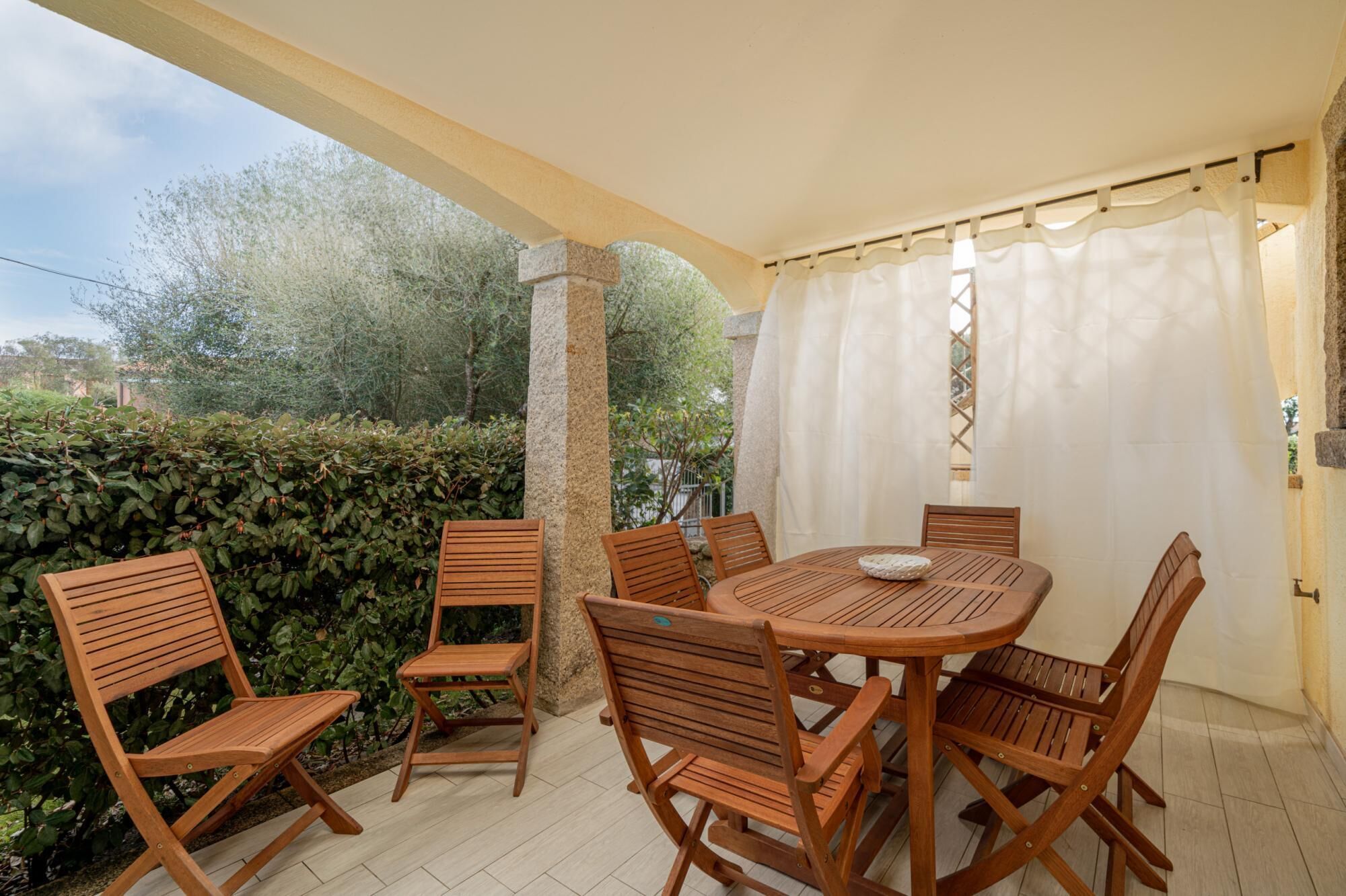 Eine überdachte Terrasse mit einem runden Holztisch und sechs Stühlen, umgeben von einer Hecke und Bäumen.