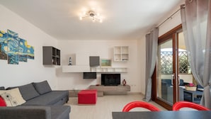 A modern living room with a grey sofa, red ottoman, and a dining table. There is a fireplace, a TV mounted on the wall, and a ceiling light fixture. - Casa Delle Ginestre by Wonderful Italy (Olbia)