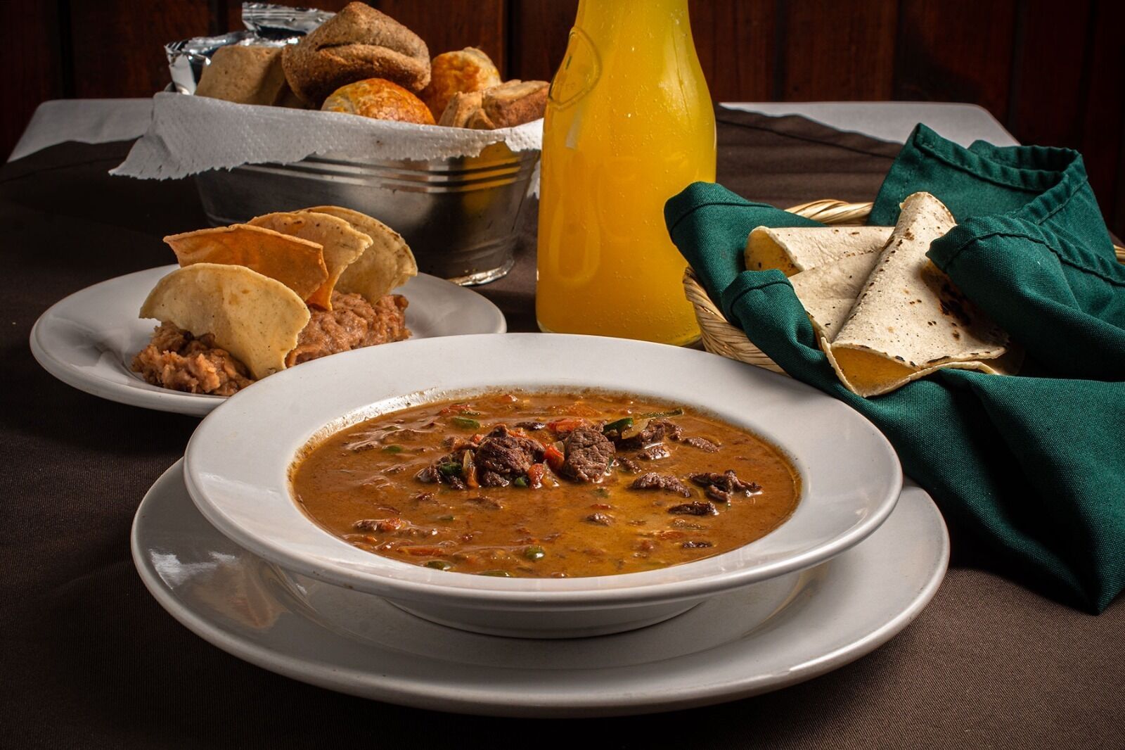 Een kom stoofpot met vlees en groenten, vergezeld van tortilla's en een fles sap op een tafel.